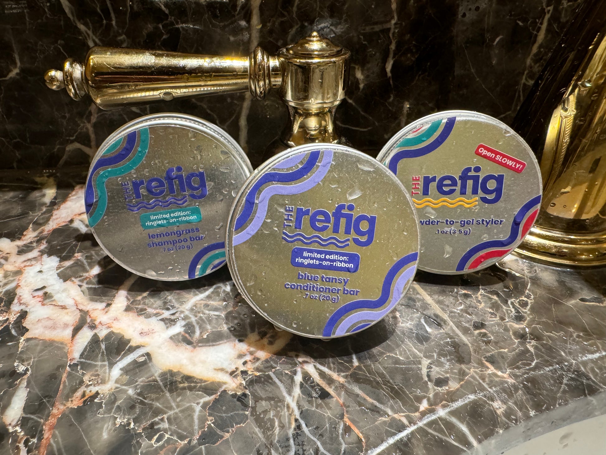 Wash-and-Go Starter Kit. Shampoo bar tin, conditioner bar tin and powder-to-gel styler tin on sink. Tins are wet to demonstrate the hydrating aspects of product.