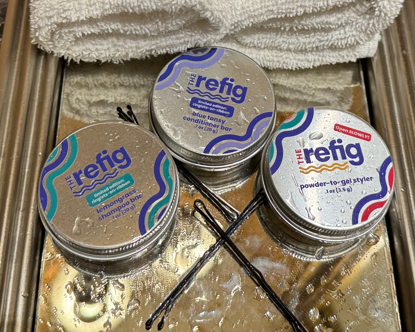 Wash-and-Go Starter Kit. Shampoo bar tin, conditioner bar tin and powder-to-gel styler tin on tray next to hair pins and towels. Tins are wet to demonstrate the hydrating aspects of product.